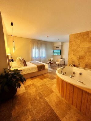 Habitación de hotel moderna con una cama grande, una zona de estar con televisor y un baño separado con bañera.