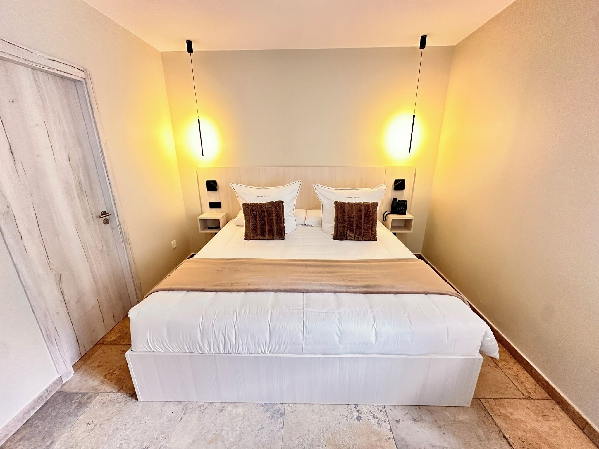 Una habitación de hotel con una cama grande, dos mesitas de noche, una lámpara de noche, una puerta y dos luces fijadas a la pared.