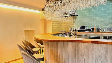 Eine Bar mit Holztresen, zwei Barhocker, einer Pendelleuchte und einer Ablage mit Flaschen und Gläsern.