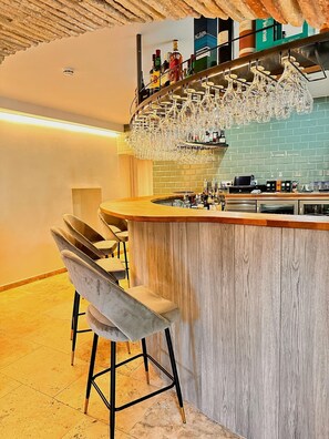 A bar area with a wooden bar counter, two bar stools, a hanging light fixture, and a shelf with bottles and glasses. - Boutique Hotel Comtal Empuries (Castello d'Empuries)