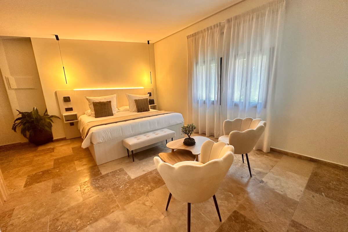 Habitació d'hotel moderna amb un llit gran, taules de nit, una banca, dues cadires i una taula petita.