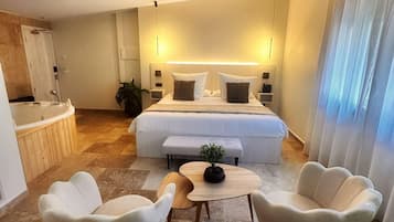 Une chambre d’hôtel moderne avec un grand lit, des tables de chevet, une petite table avec une plante, deux fauteuils blancs et une salle de bain équipée d’une baignoire et d’une douche.