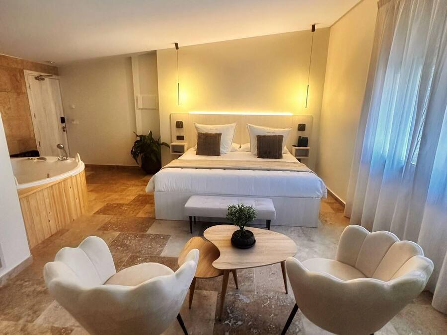 Ein modernes Hotelzimmer mit einem großen Bett, Nachttischen, einem kleinen Tisch mit Pflanze, zwei weißen Sesseln und einem Badezimmer mit Badewanne und Dusche.