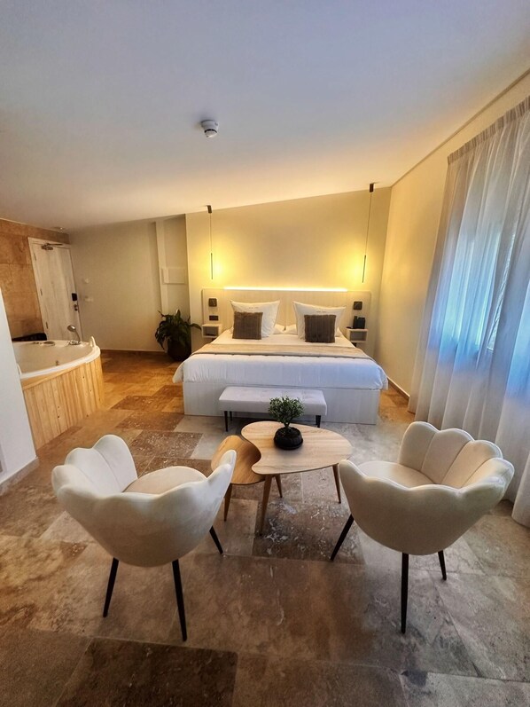 Una habitación de hotel moderna con una cama grande, mesitas de noche, una pequeña mesa con una planta, dos sillones blancos y un baño con bañera y ducha.