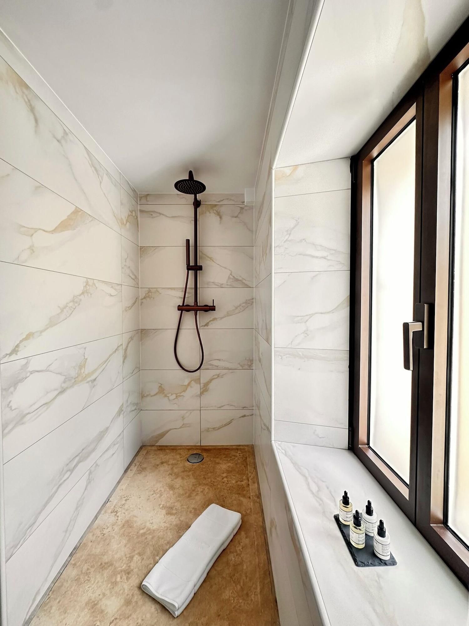A modern bathroom with marble walls, a shower with a handheld head, a towel on the floor, and toiletries on a shelf by the window.