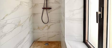 Un bagno moderno con pareti in marmo, una doccia con soffione a mano, un asciugamano sul pavimento e articoli da toilette su uno scaffale vicino alla finestra.