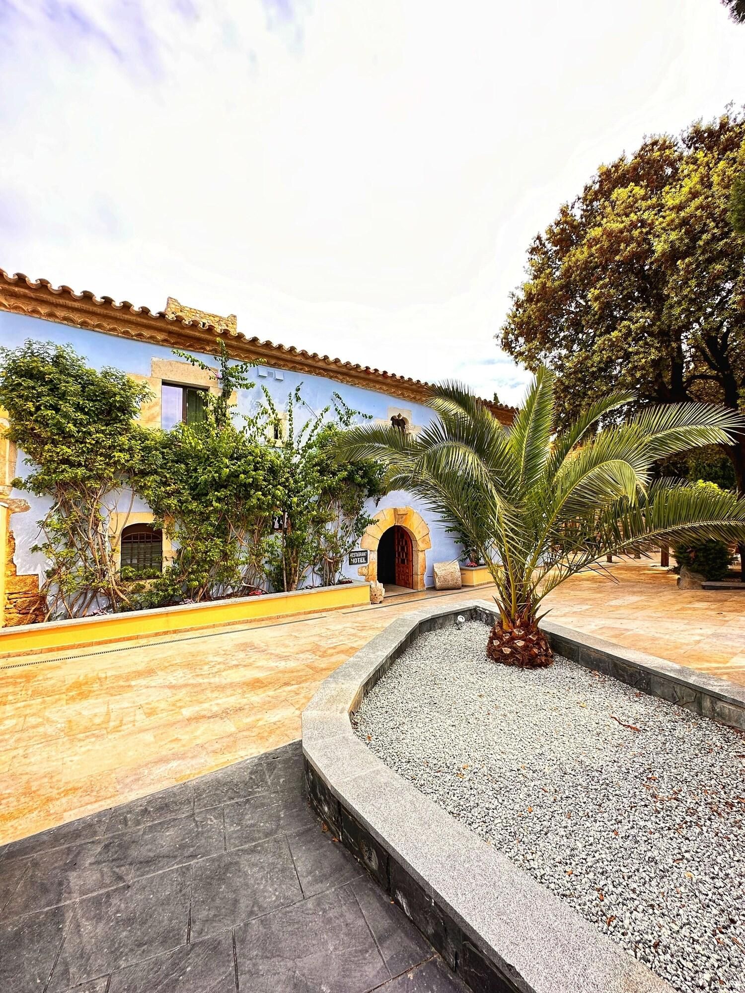 A courtyard with a building featuring a tiled roof and arched doorways, surrounded by greenery and a palm tree.