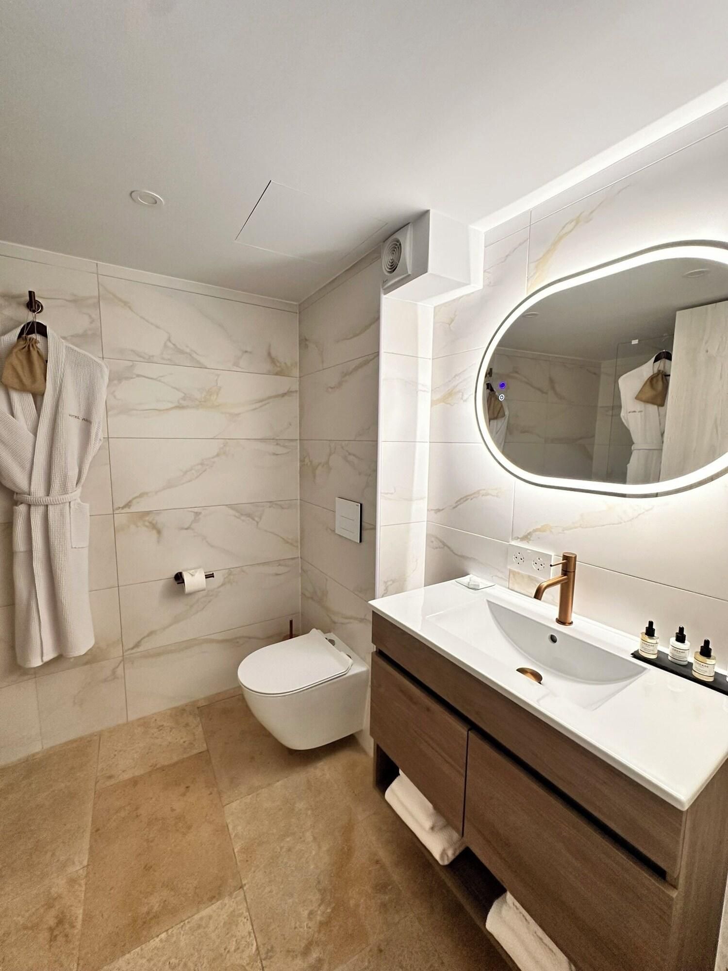 Un baño moderno con salpicadero de mármol, un espejo ovalado grande, un inodoro blanco y un tocador de madera con lavabo.
