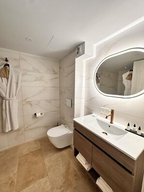 Un baño moderno con salpicadero de mármol, un espejo ovalado grande, un inodoro blanco y un tocador de madera con lavabo.