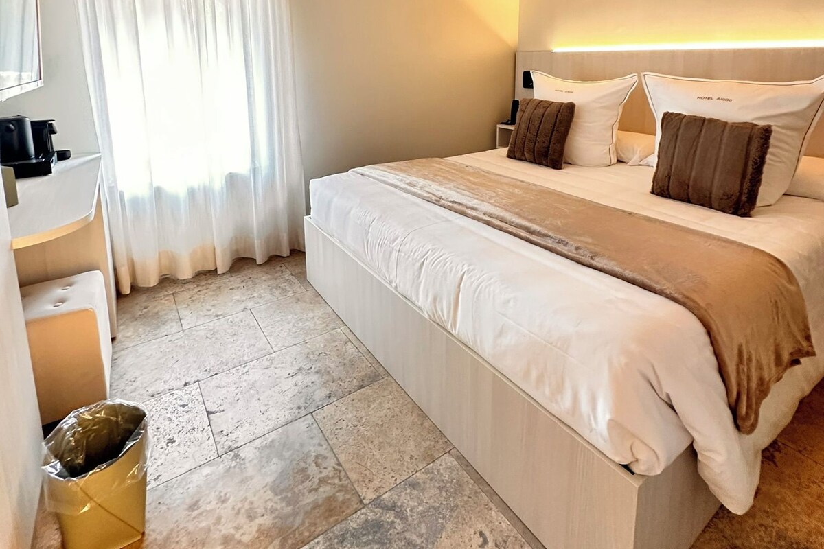 Una habitación de hotel moderna con una cama grande, una mesita de noche, un espejo y una ventana con cortinas.