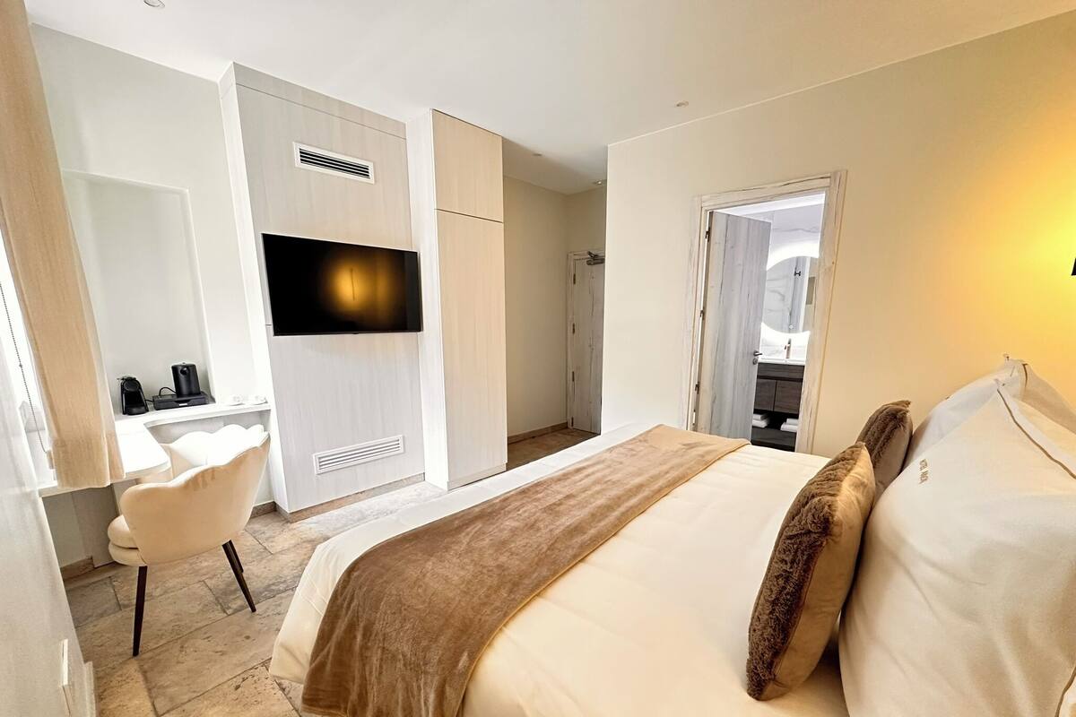 A hotel room with a bed, a desk, a chair, a TV, and a bathroom visible through an open door.