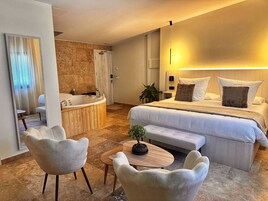 Una habitación de hotel moderna con una cama grande, una zona de estar con dos sillones, una mesita, una bañera e una planta en maceta.