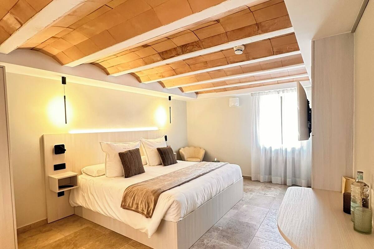 A bedroom with a large bed, a bedside table, a desk, and a ceiling with wooden beams.