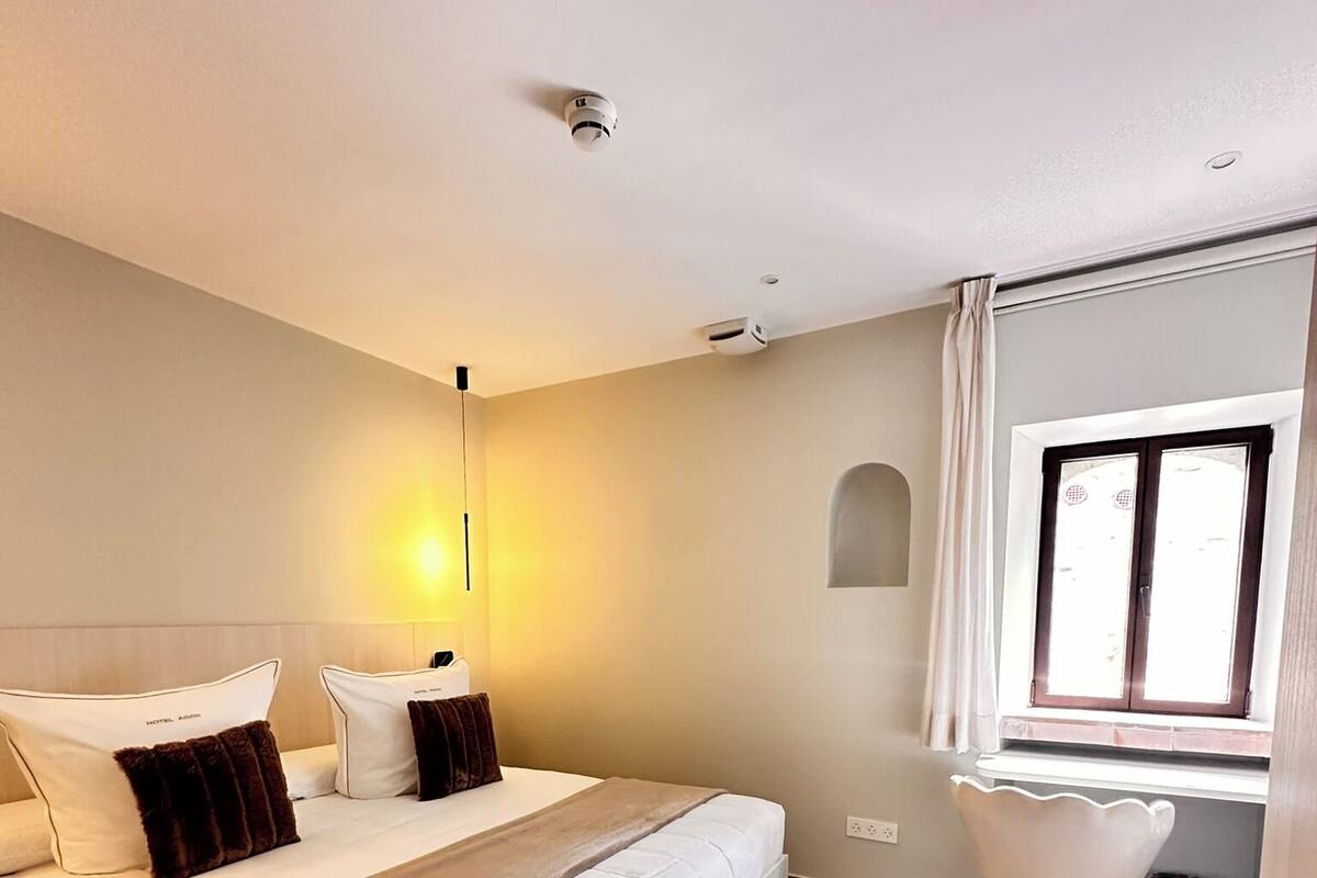 A hotel room with a large bed, a bedside table, a chair, a window, and a wall-mounted air conditioning unit.