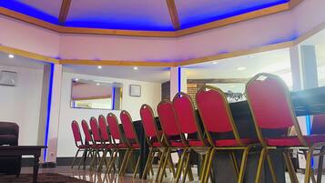 Una sala de conferencias con un techo que presenta un diseño geométrico y una lámpara central. Sillas rojas dispuestas en filas, y una mesa visible en primer plano.