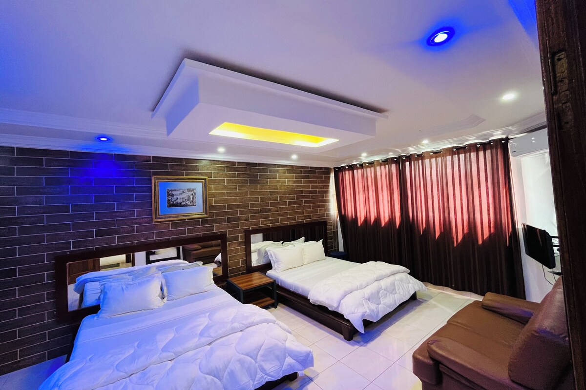 A hotel room with two beds, a small sofa, a brick-patterned wall, and a picture frame.