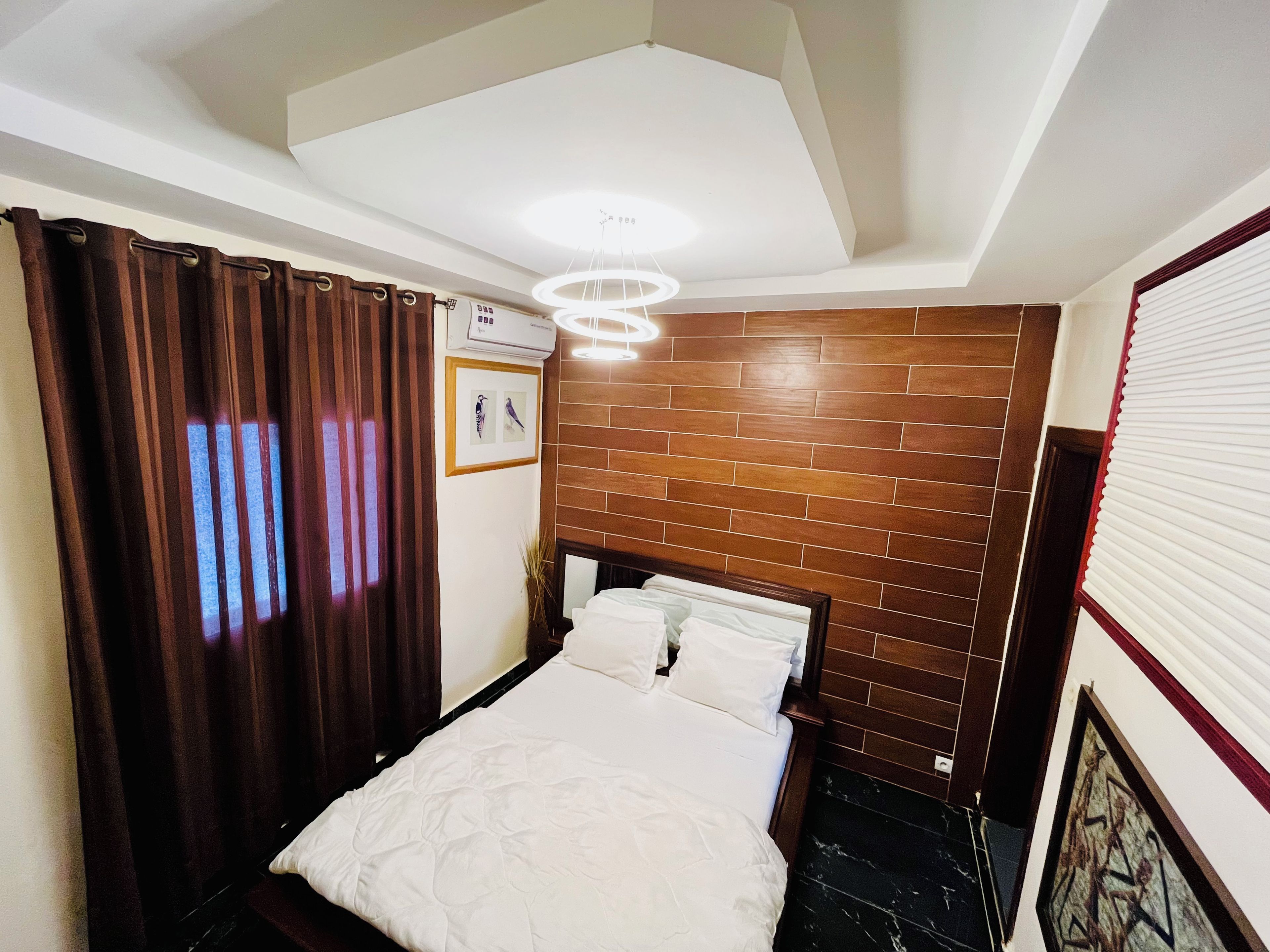 A hotel room with a bed, a wooden headboard, a wall-mounted air conditioner, a ceiling light, and a framed picture on the wall.