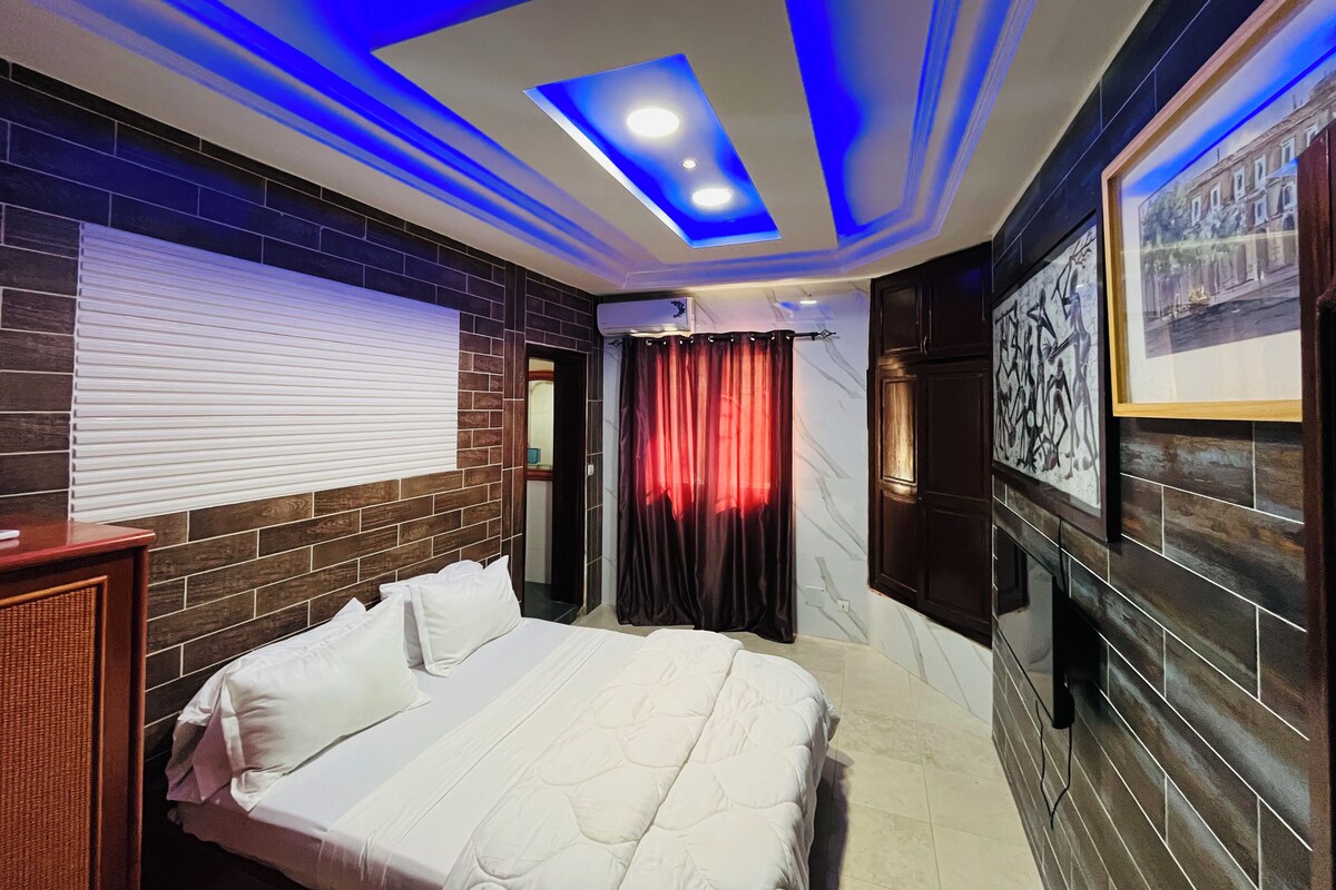A hotel room with a bed, a window with blinds, a wall-mounted air conditioner, and a ceiling with blue lighting.