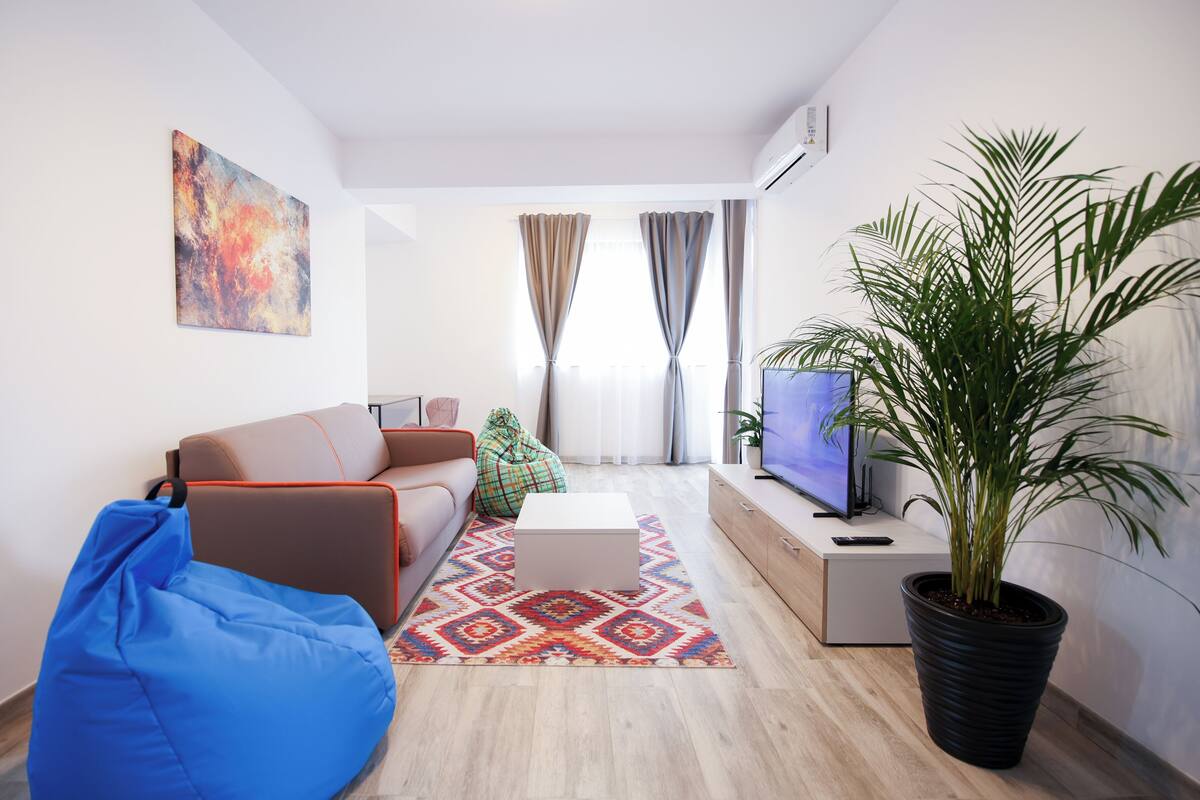Una sala de estar moderna con un sofá, un sillón de peluche azul, una mesa de centro, un televisor, una planta en maceta y una alfombra colorida.