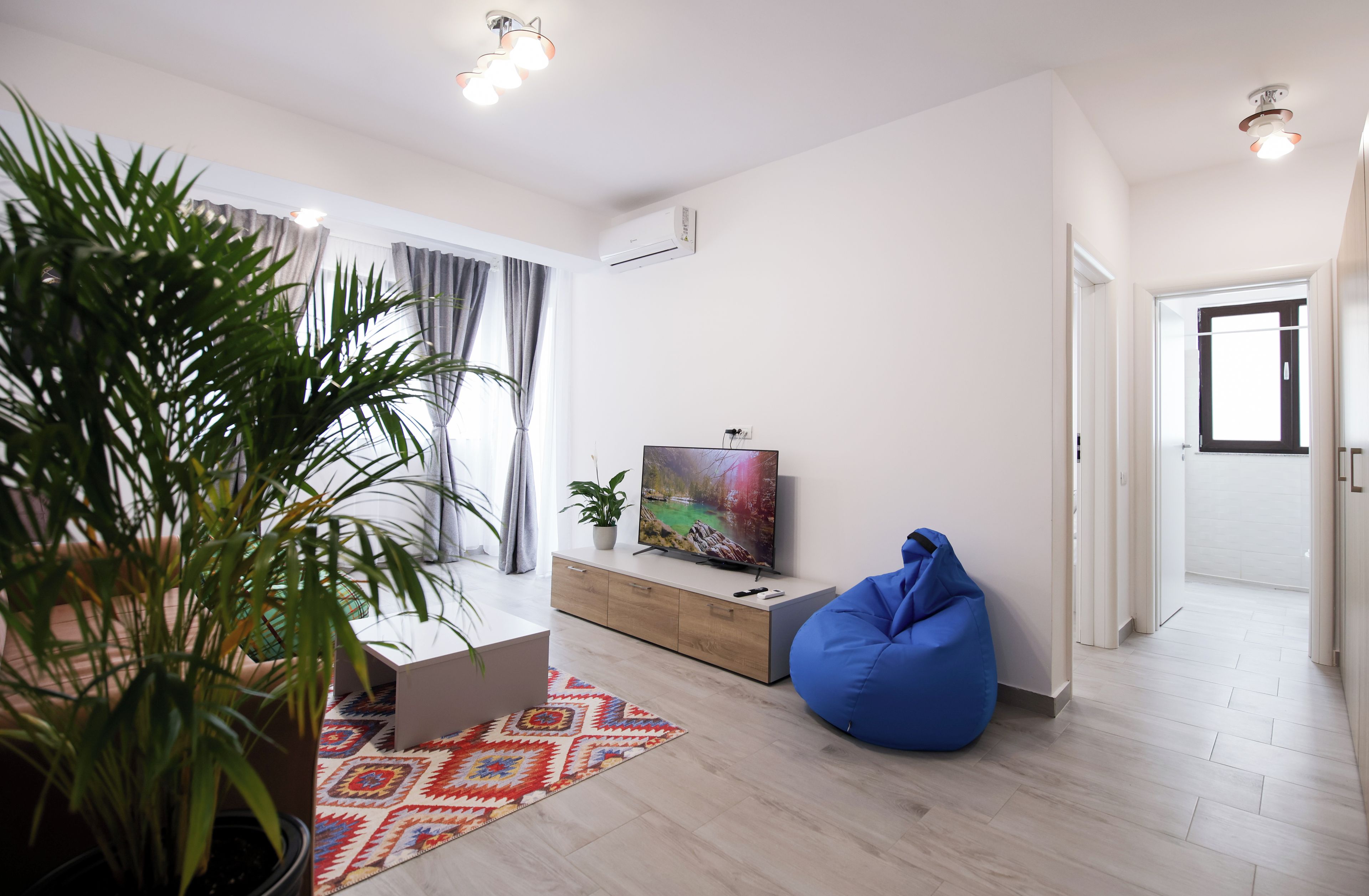 Uma sala de estar moderna com uma janela ampla, um puff azul, uma TV de tela plana e um tapete estampado.