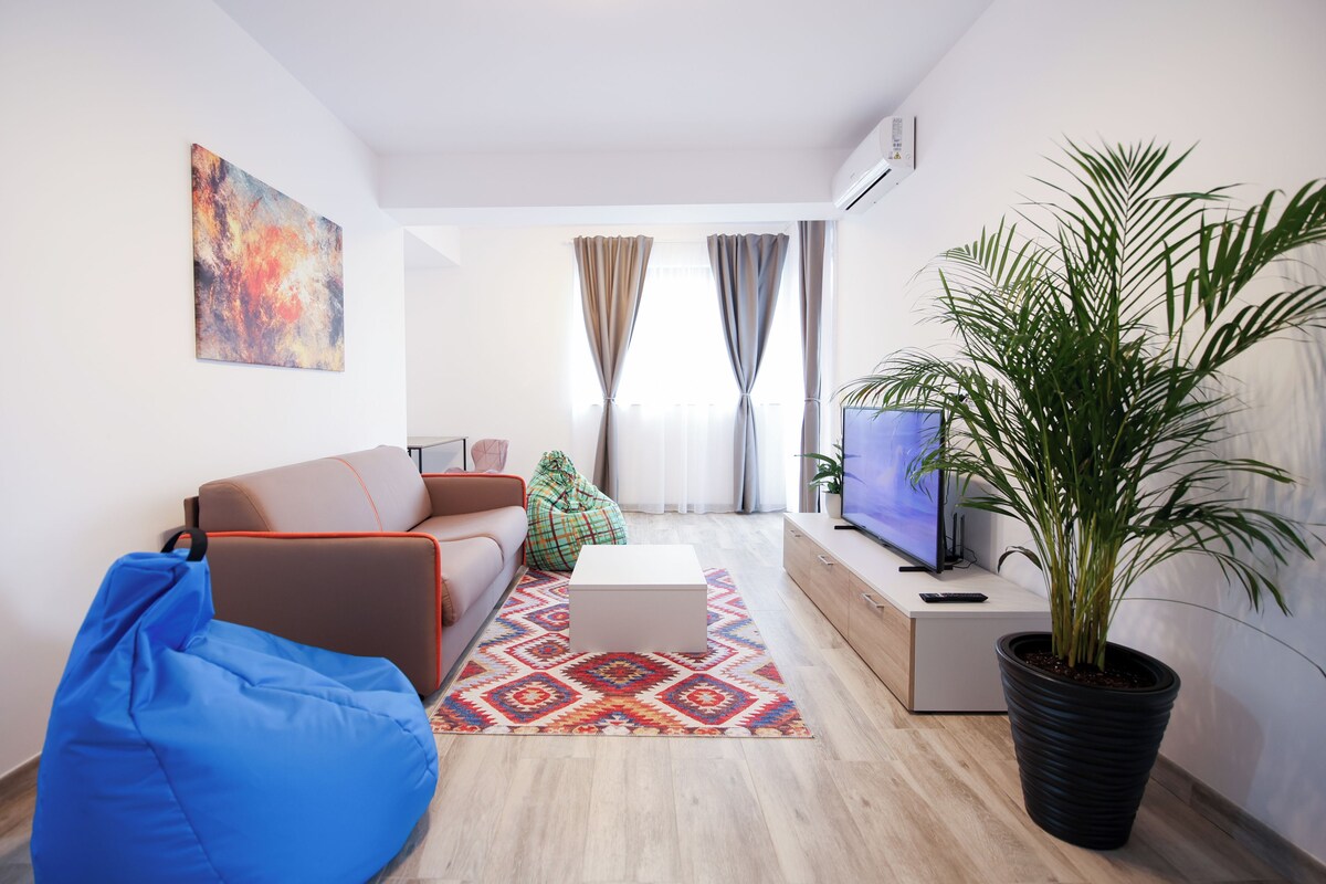A modern living room with a sofa, a blue bean bag, a coffee table, a TV, a potted plant, and a colorful rug.