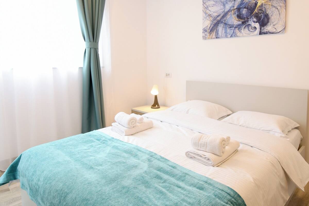 Una cama bien hecha con un edredón azul verdoso, almohadas blancas y una lámpara de noche.