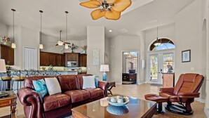 A spacious living room with a leather sofa, a recliner, and a coffee table. There is a kitchen in the background with wooden cabinets and a dining area. - Intervillas Florida - Villa Royal Sereni Tee (Cape Coral)