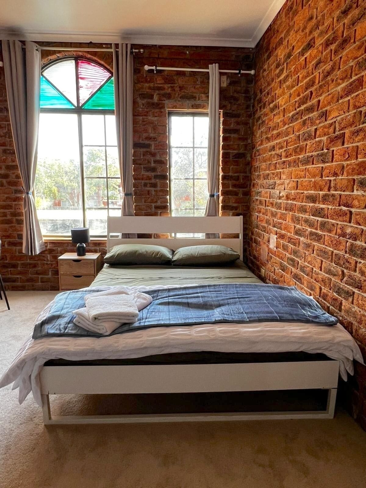 A bedroom with a brick wall, a bed with a white headboard, two windows with curtains, and a nightstand with a lamp.