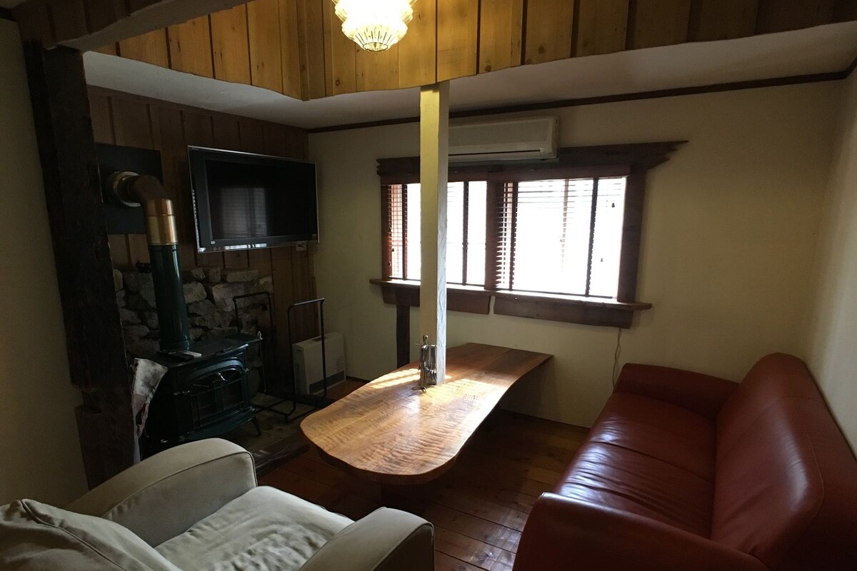 A cozy room with a wooden ceiling, a stone wall, a fireplace, a television, a wooden table, a sofa, and a window with blinds.