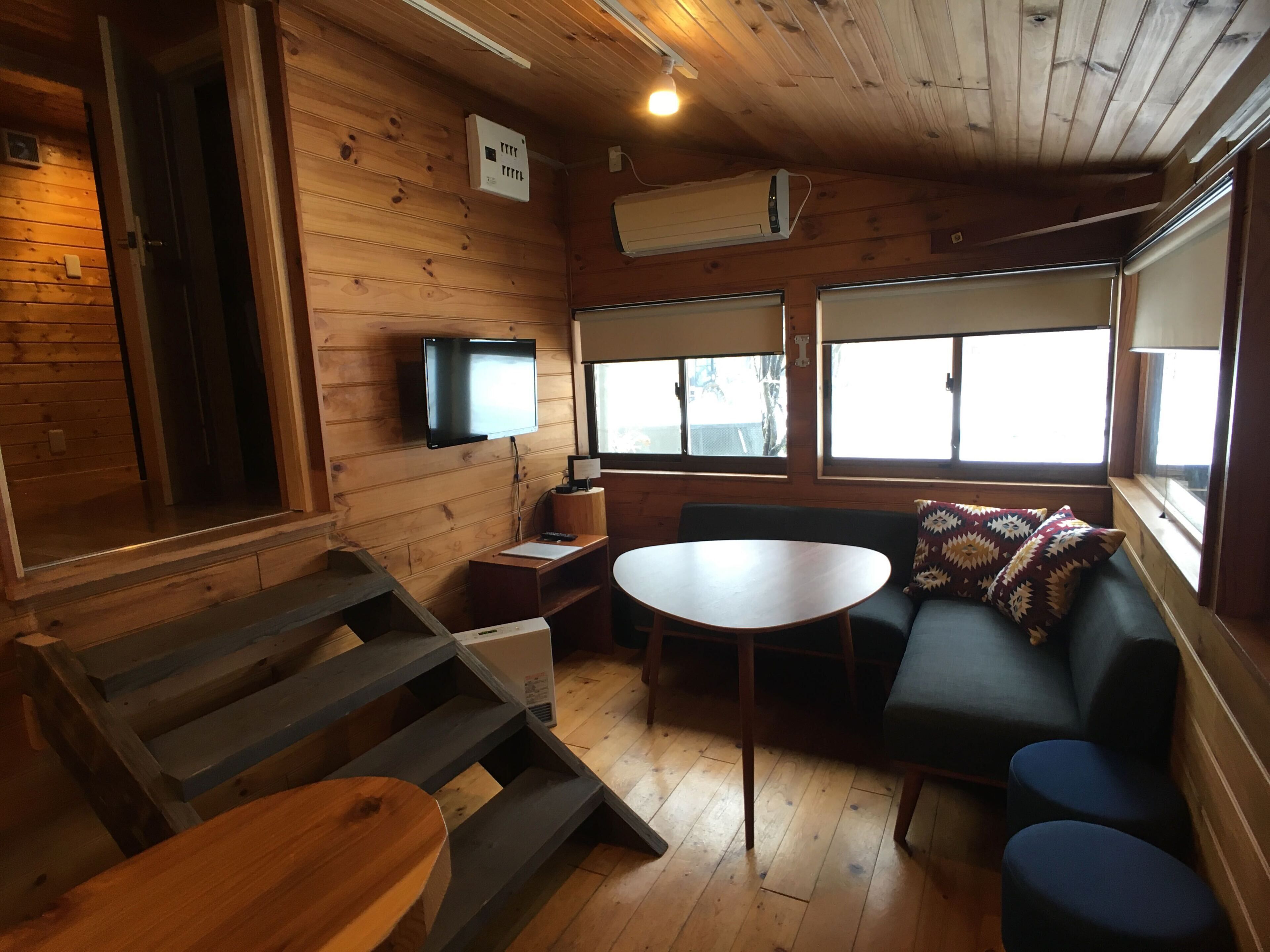 A cozy cabin interior with a wooden floor, a sofa, a table, a TV mounted on the wall, and large windows.