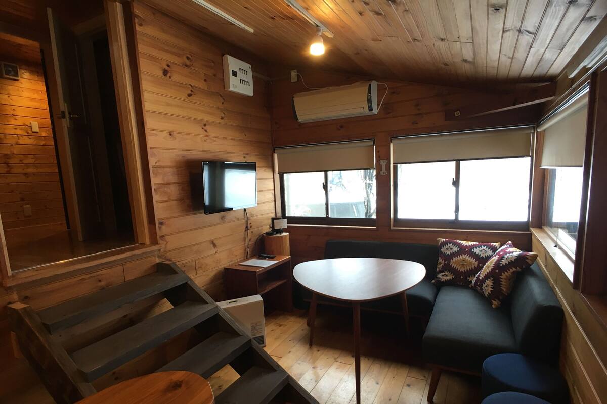 A cozy cabin interior with a wooden floor, a sofa, a table, a TV mounted on the wall, and large windows.