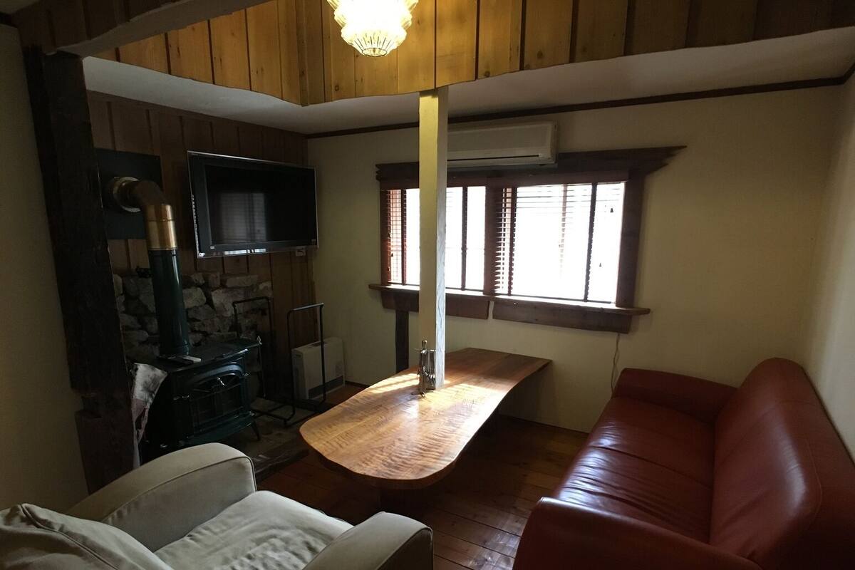 A cozy room with a wooden ceiling, a stone wall, a fireplace, a television, a wooden table, a sofa, and a window with blinds.