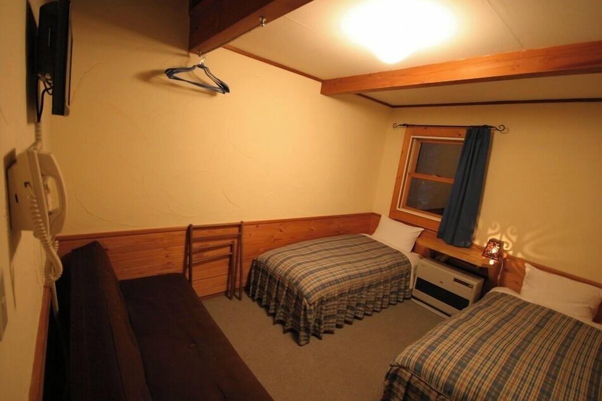 A small, cozy room with two beds, a wooden bench, a desk with a lamp, and a window with curtains.