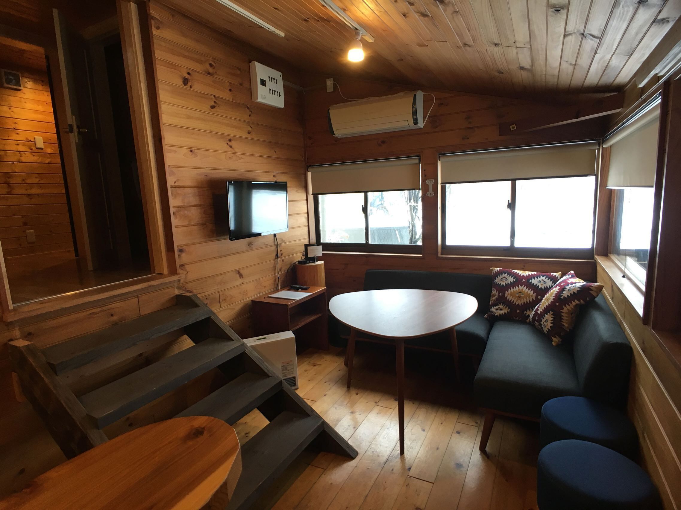 A cozy cabin interior with a wooden floor, a sofa, a round table, a TV mounted on the wall, and large windows.