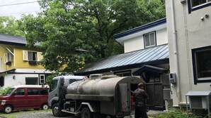 A building with a metal roof, a parked red car, and a large industrial tank truck. - Hakuba Himekawa Onsen Pension Tomato (Hakuba)