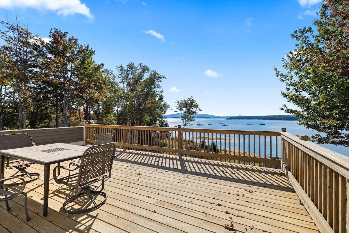 A wooden deck with a table and chairs, overlooking a body of water with boats and distant land.