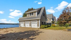 Una casa a due piani con un garage, una spiaggia sabbiosa davanti e un lago sullo sfondo.