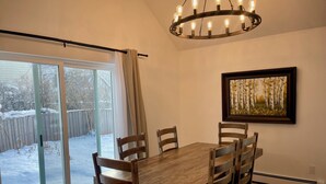 Un coin repas avec une table et des chaises en bois, un lustre, une porte-fenêtre coulissante donnant sur l’extérieur et un tableau accroché au mur.