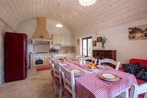 Zona pranzo con tovaglia a scacchi, sedie bianche, frigorifero rosso e cucina sullo sfondo.