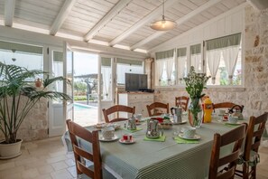 Un comedor con una mesa preparada para cuatro personas, sillas, un televisor y una planta grande en maceta.