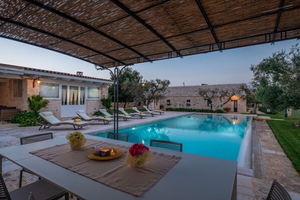 Un área junto a la piscina con sillas de descanso, una mesa con vela y flores, y un porche cubierto.