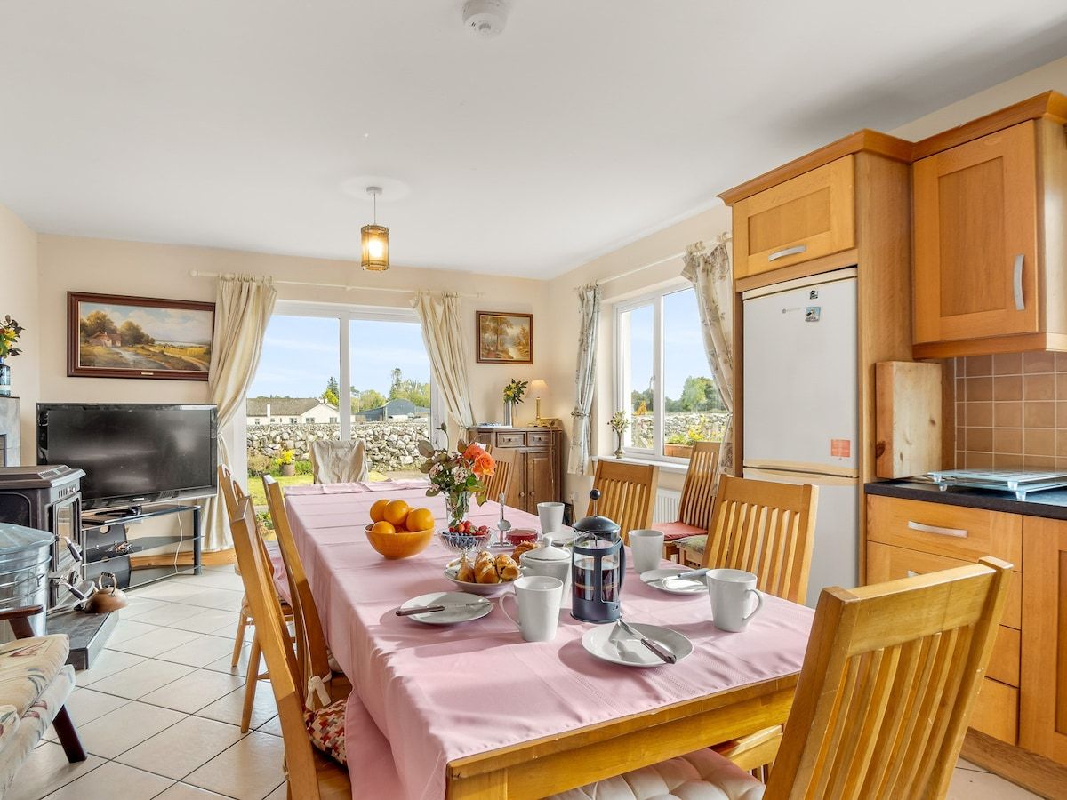 A dining area with a long table set for breakfast, a television, and a kitchen area with wooden cabinets.