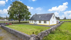 A white house with yellow trim, a stone wall, and a large tree in front.