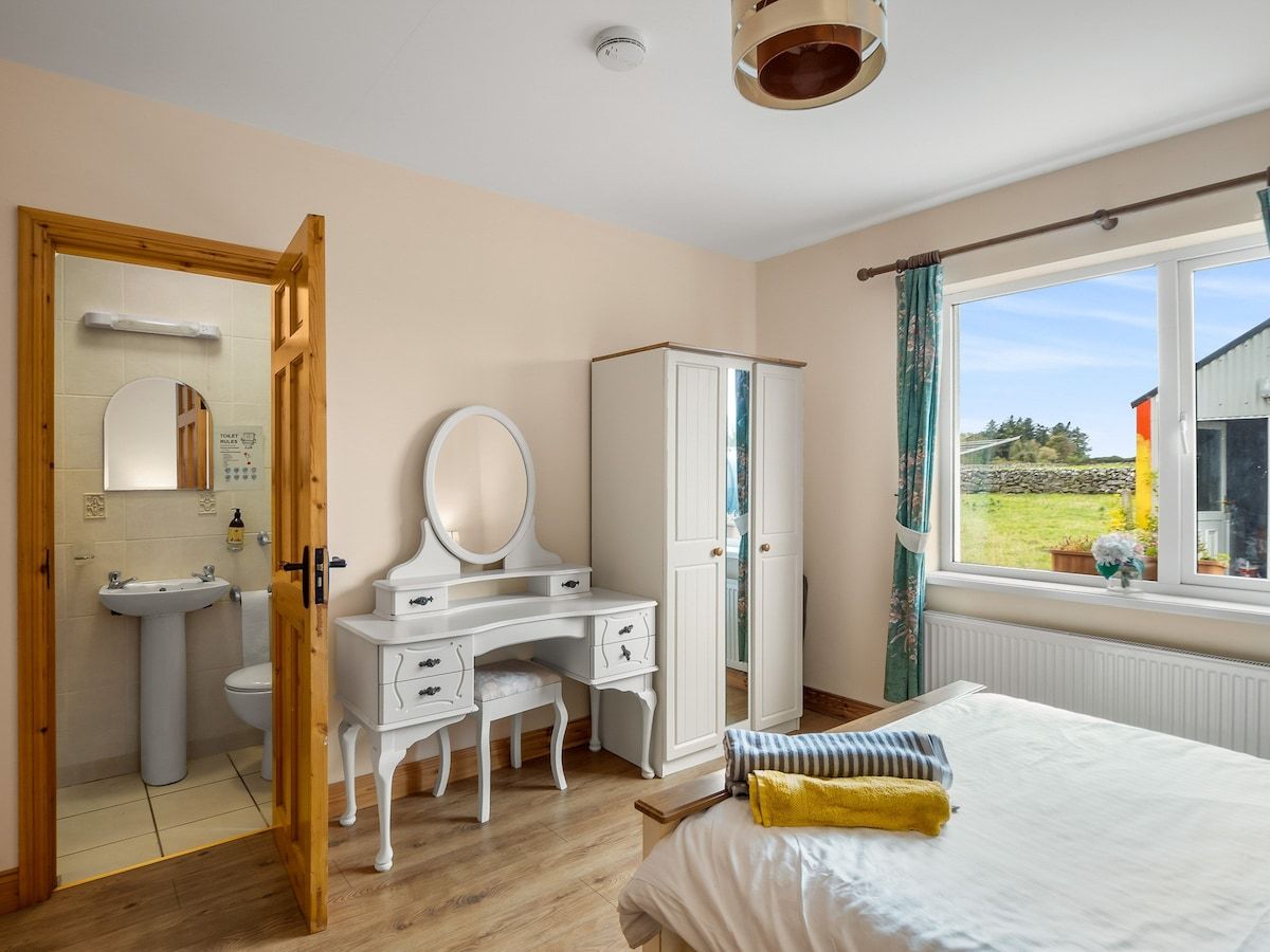 Un dormitorio con cama, cómoda, tocador con espejo, ventana con vistas al exterior y puerta que da a un baño.