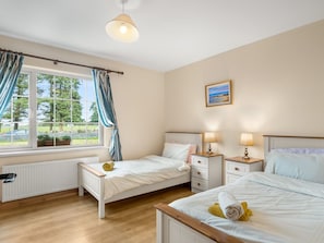 A bedroom with two single beds, a window with curtains, a radiator, and a bedside table with lamps.
