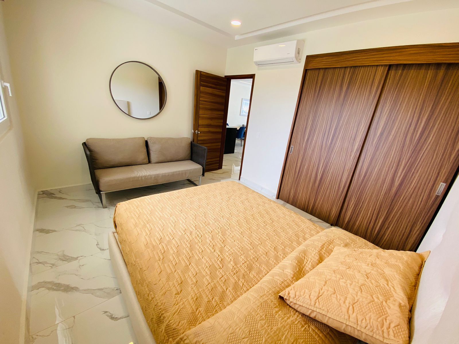 A bedroom with a bed, a sofa, a wardrobe, a mirror, and an air conditioning unit.