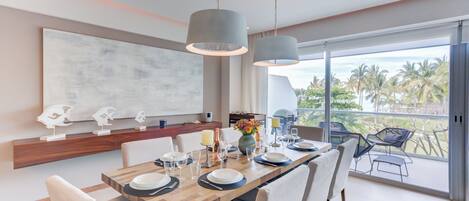 Un comedor moderno con una mesa de madera dispuesta para seis comensales, grandes lámparas colgantes y vistas a palmeras a través de puertas de vidrio amplias.