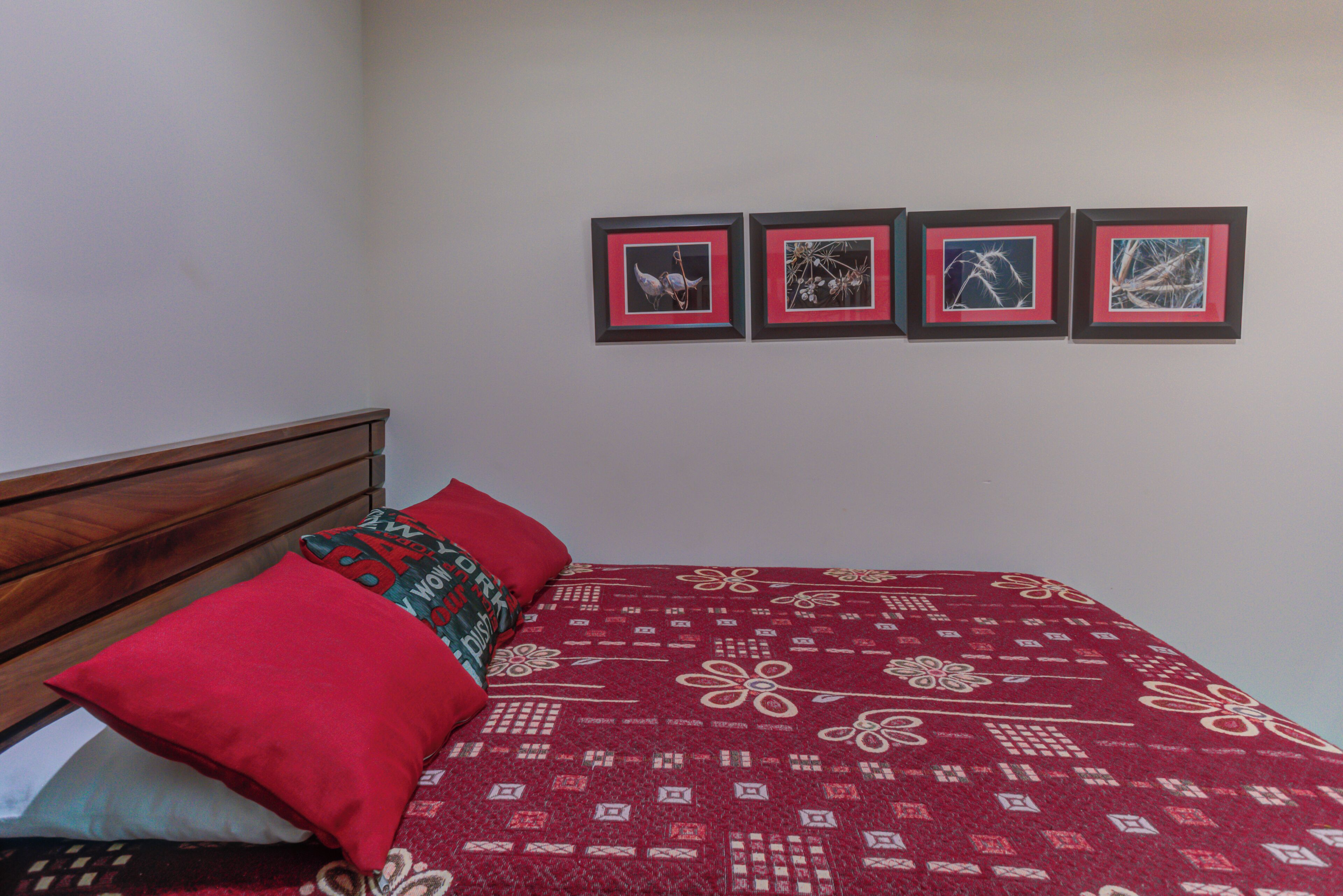 Una cama con una colcha roja estampada, un cabecero de madera y tres cuadros en la pared.