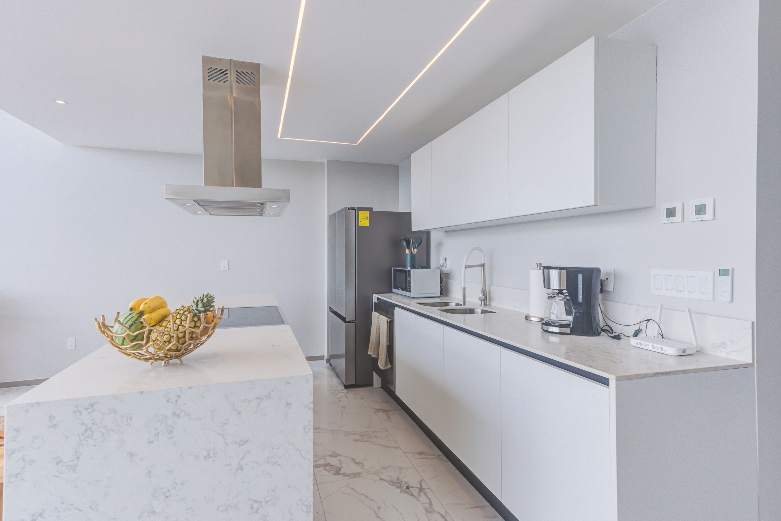 A modern kitchen with white cabinetry, a marble countertop, a stainless steel range hood, a refrigerator, a microwave, a coffee maker, and a fruit bowl.