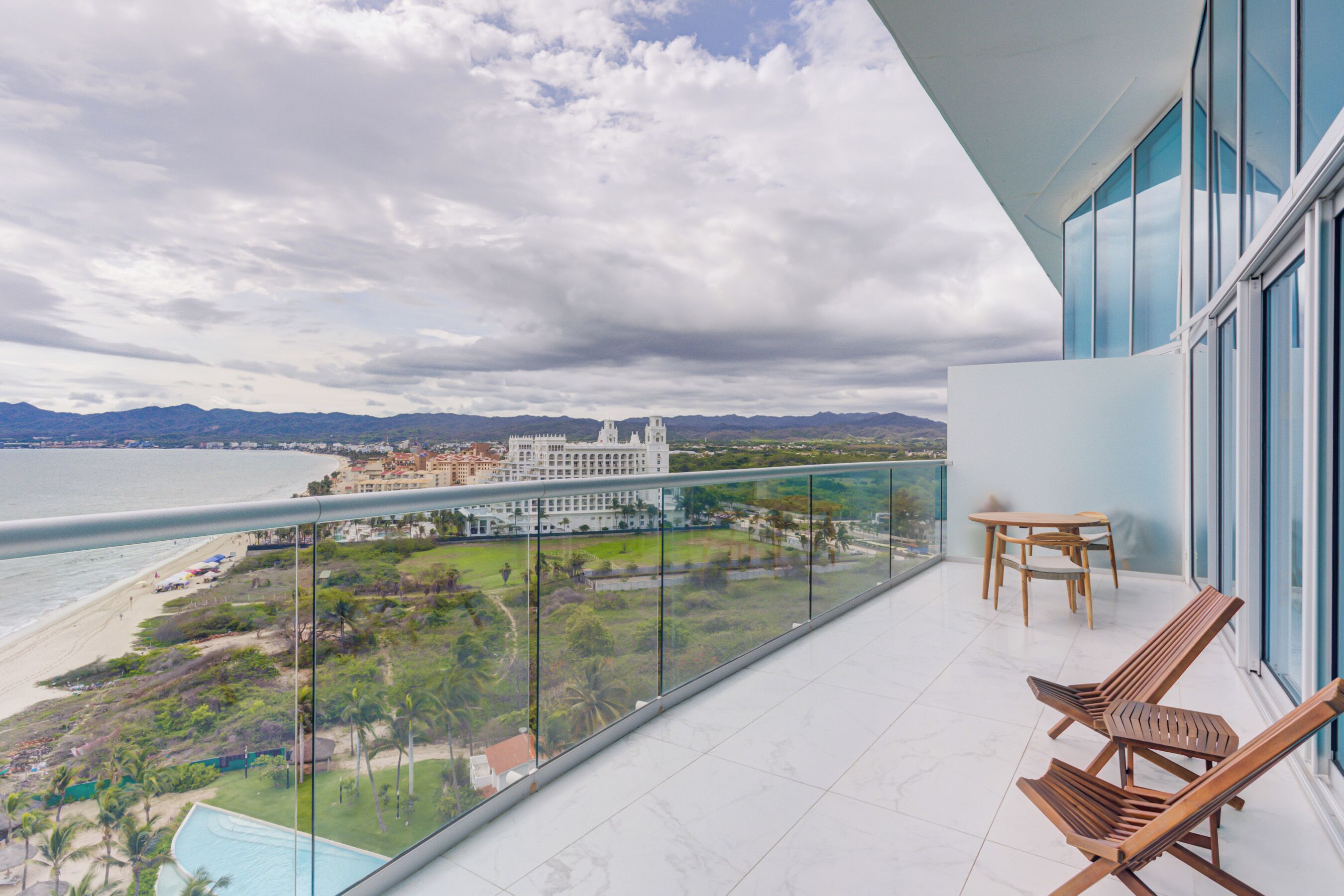 A balcony with a view of a beach, a pool, and a large building in the distance.